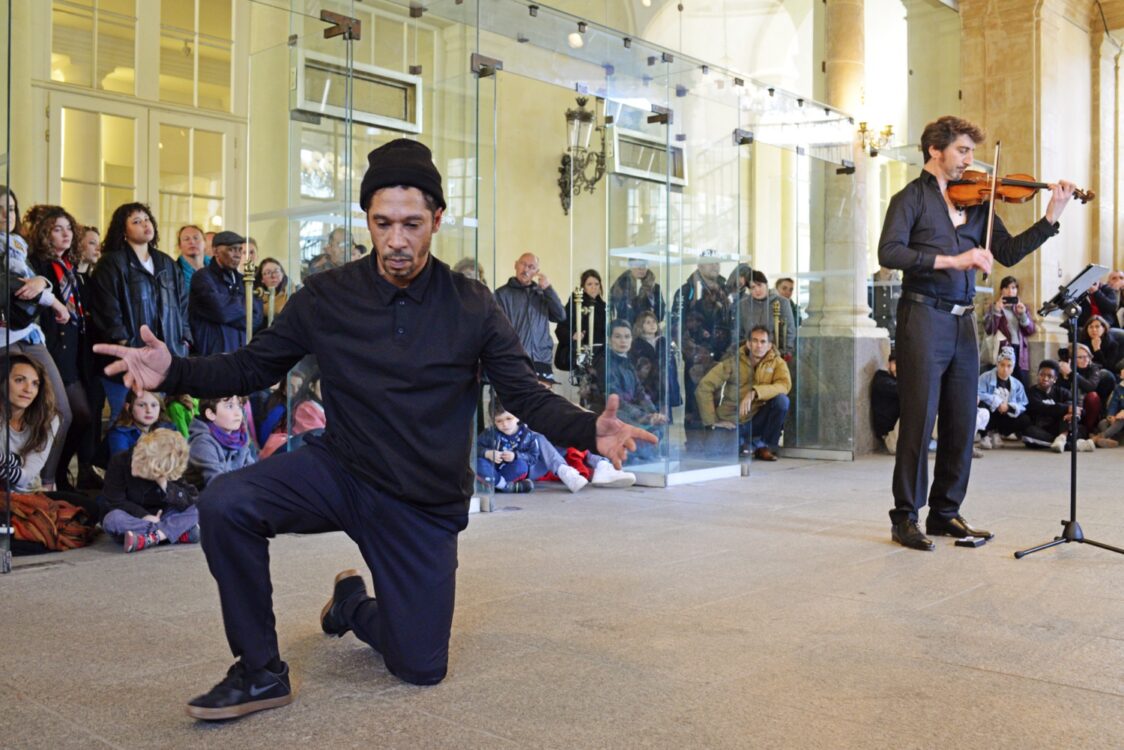 Danseur en mouvement, violoniste à côté avec spectateur en fond.