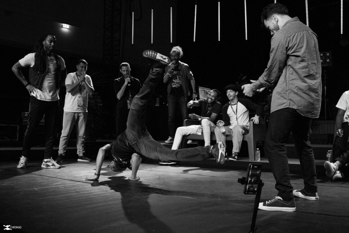 Image en noir et blanc. Un danseur en mouvement a sol au centre. Des danseurs qui le regardent autour.