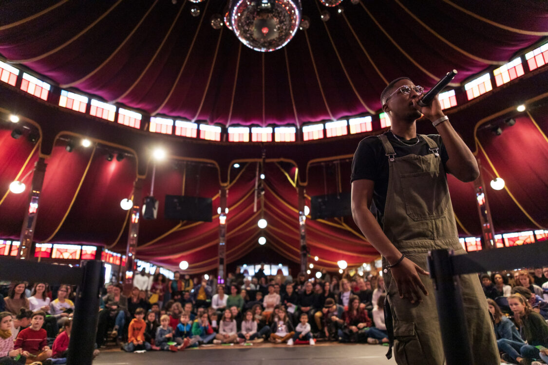 Personne tenant un micro en premier plan. Derrière une scène avec des enfants et un public par terre ou debout.