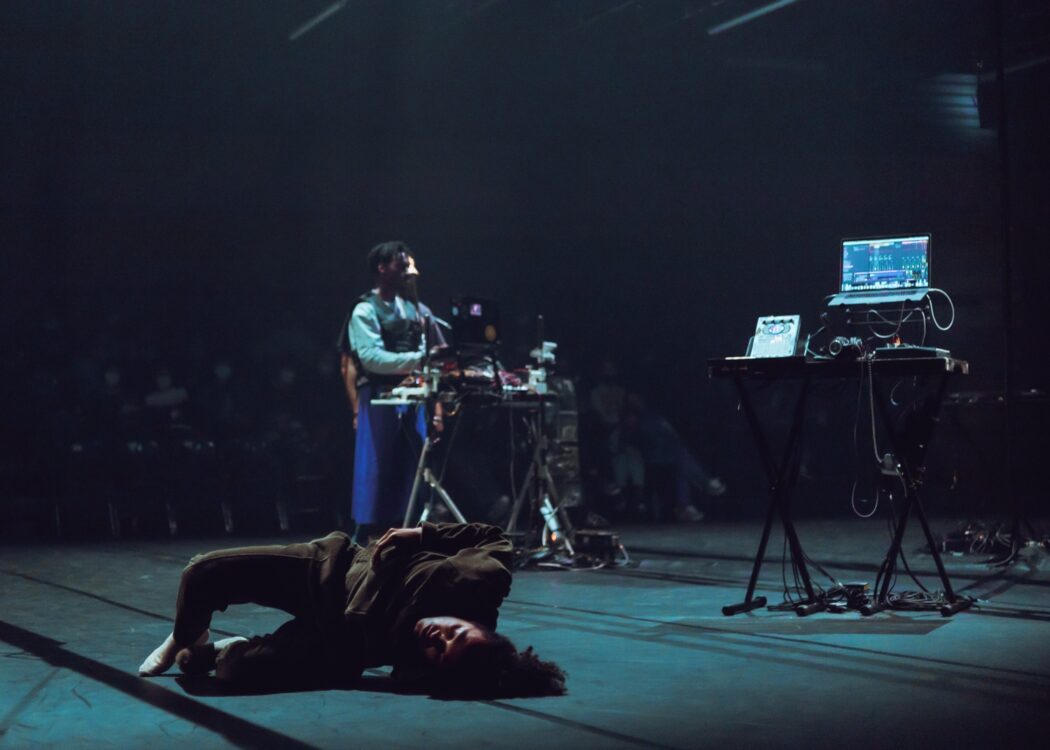 Danseur en mouvement au premier plan avec un musicien et le public en fond durant le spectacle Earthbound.