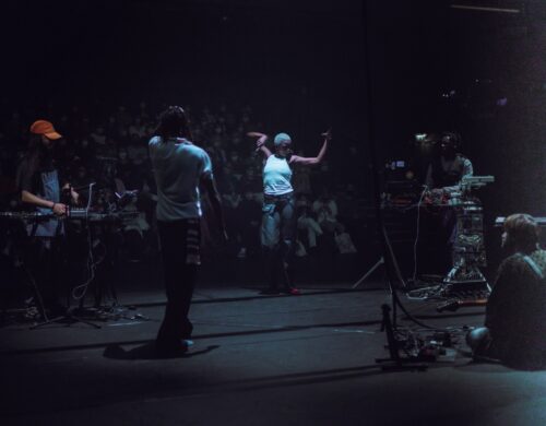 Danseuse en mouvement au centre, musiciens jouant et spectateurs en fond.