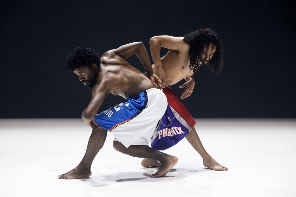 Deux danseurs en mouvement, leurs corps sont côte à côte un qui regarde droit devant lui.