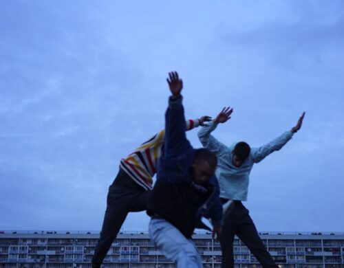 Trois danseurs en mouvement. Fond est un immeuble en large.