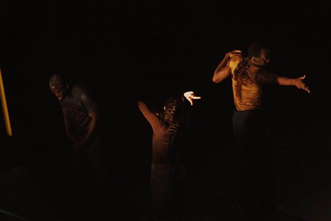 Trois danseurs sur scène en mouvement. Lumière jaune sur eux, fond noir.