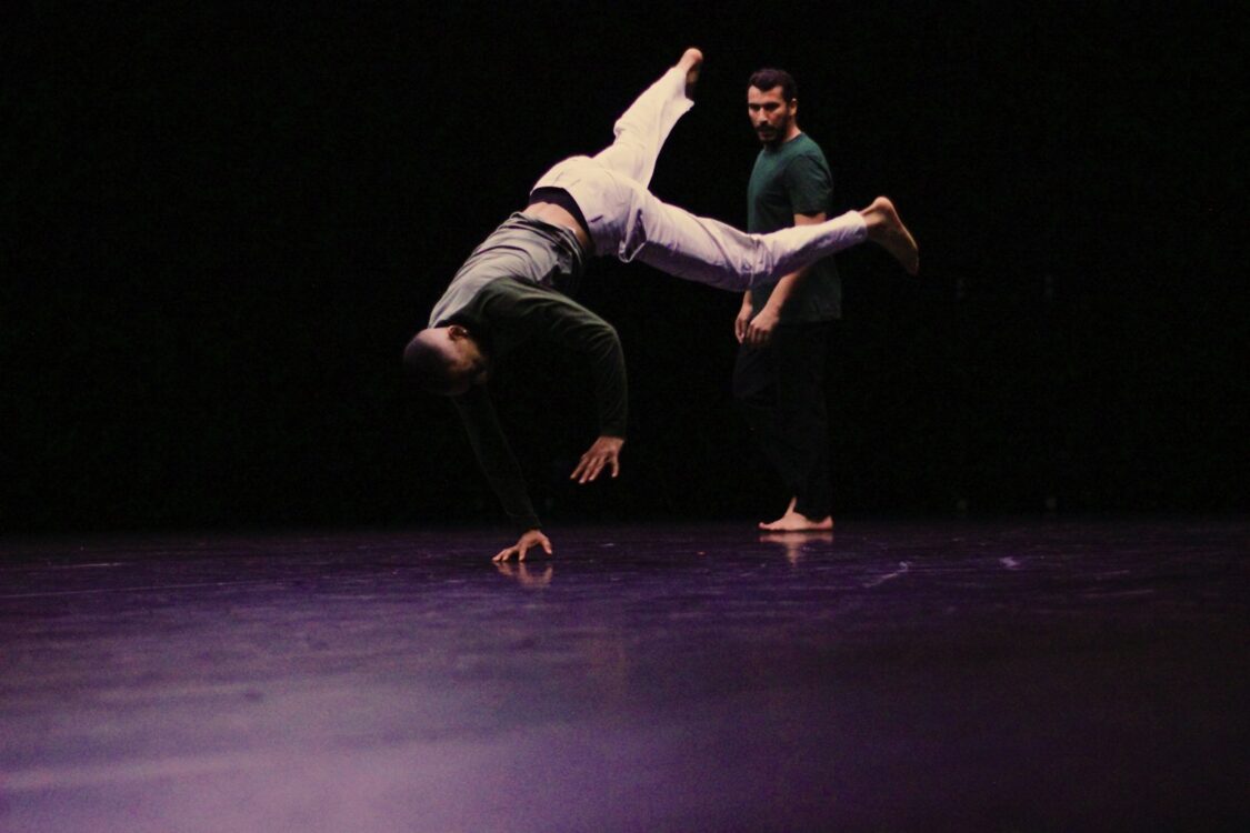 Un danseur en mouvement de breakdance, une main au sol, jambes en l'air. Un autre danseur dans le fond qui le regarde.