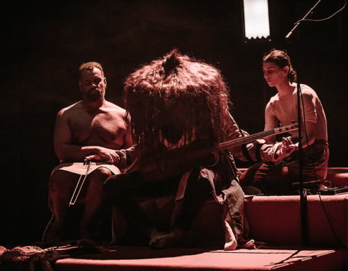 Trois personnes côte à côte. La personne de gauche a le regard perdu au loin. La personne au centre joue d'un instrument et porte un grand chapeau pointu fait de cheveux. La personne à droite regarde vers le bas.
