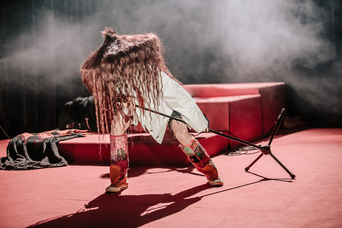 Personne tenant un micro et portant un grand pointu fait de cheveux. Le visage est caché et le corps est penchée en avant, jambes pliées.